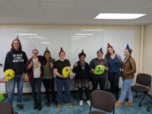 eight people pose for a picture. all are wearing party hats and there are two balloons with radioactive symbols on them.